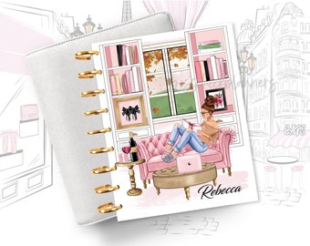 May include: A pink and white planner with gold rings. The cover features a woman sitting on a pink sofa reading a book. The background is a pink and white bookshelf with a window behind it. The text "Rebecca" is written in black at the bottom of the planner.