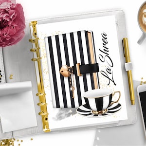 May include: A white and black striped notebook with a gold lock and key charm. The notebook is open to a page with gold glitter and the text "La Shree". A black and white teacup and saucer are on the page.