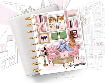 May include: A pink and white planner with a gold coil binding. The cover features a woman sitting on a pink couch reading a book. The background is a drawing of a Parisian street scene with a cafe and a building with a balcony. The planner has the name "Jacqueline" printed on the cover.