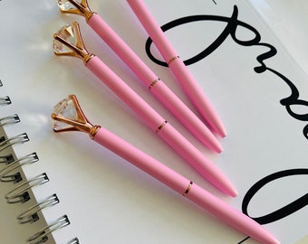 May include: Four pink pens with gold accents and clear diamond-shaped tops. The pens are arranged on a white notebook with the word "2022" written at the top.