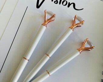 May include: Three white pens with gold diamond-shaped tops. The pens are arranged in a triangular shape on a white surface. The text "Vision" is written in black ink on the surface.