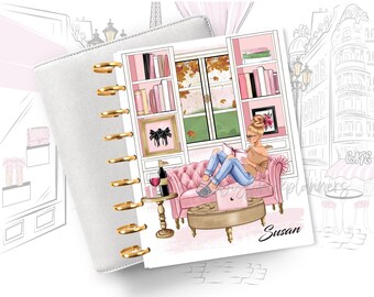 May include: A planner cover with a pink and white design featuring a woman sitting on a pink couch reading a book. The design includes a bookshelf, a window with fall leaves, a glass of wine, and a laptop. The text "Susan" is at the bottom of the cover.