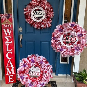 May include: Three red, white, and purple wreaths with the Greek letters Delta Sigma Theta and the words "Delta Sigma Theta Sorority, Inc. 1913 All of My Love" and "Devastating Diva" on them. The wreaths are hanging on a blue door with a red and white welcome sign.