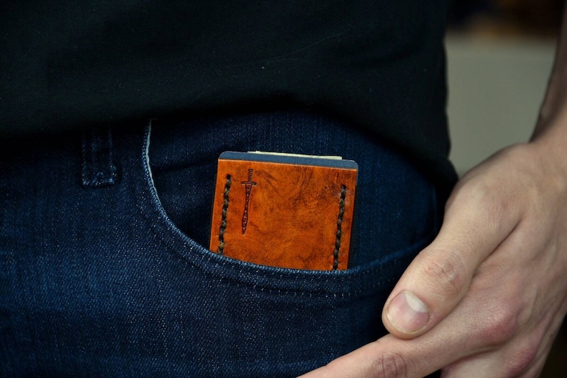 Op de afbeelding: Een bruine leren portemonnee met een gestikt ontwerp en een klein zwaardsymbool. De portemonnee zit in een blauwe spijkerbroekzak.