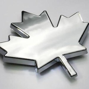 May include: A shiny, chrome-finished maple leaf emblem. The leaf has a detailed, multi-pointed shape with a stem. The surface reflects light, creating a bright, metallic appearance. The emblem is likely for automotive or decorative use.