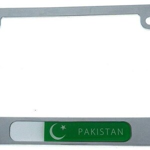 May include: A silver-colored license plate frame with a green and white Pakistan emblem. The frame has four mounting holes and a rectangular section with the word "PAKISTAN".