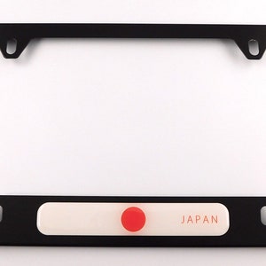 May include: Black license plate frame with a white and red accent. The accent features the word "JAPAN" in a red font and a red circle. The frame is rectangular with mounting holes.