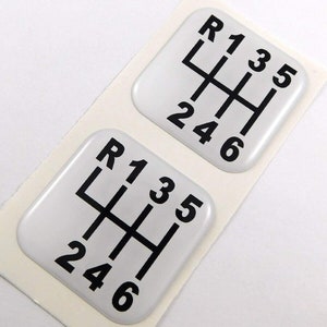 May include: Two white gear shift stickers with black lettering. The stickers show the gear shift pattern with the numbers R, 1, 2, 3, 4, 5, and 6. The stickers are on a white background.