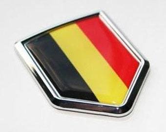 May include: A rectangular emblem with a chrome border, featuring the colors of the Belgian flag: black, yellow, and red. The emblem has a glossy finish and a slightly raised surface, suggesting a three-dimensional design.