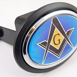 May include: Black and silver hitch cover with a blue oval emblem featuring a Masonic symbol. The emblem has a gold "G" in the center, surrounded by a square and compass design. The hitch cover is designed to fit a standard 2-inch receiver.