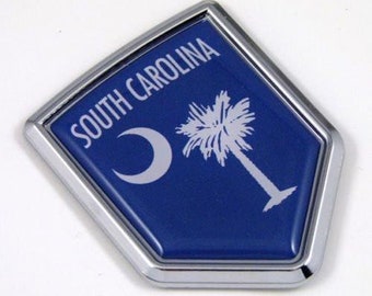 May include: A diamond-shaped emblem featuring the South Carolina state flag design. The emblem has a blue background with a white crescent moon and a palmetto tree. The text "SOUTH CAROLINA" is displayed in white. The emblem is framed with a silver border.