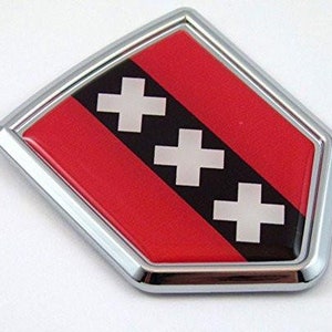 May include: A chrome-framed emblem featuring the Amsterdam coat of arms. The design includes three white crosses on a black stripe, set against a red background. The emblem has a shiny, reflective finish.