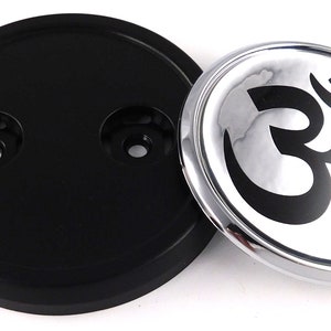 May include: A black plastic base with two holes and a round, silver-rimmed emblem. The emblem features a black Om symbol on a white and gray background. The items are likely decorative.