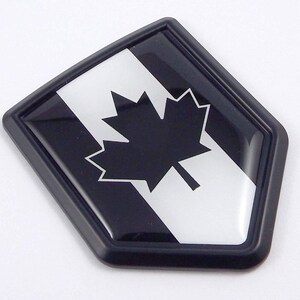 May include: A black and white emblem with a maple leaf design, set against a white background and a black border. The emblem has a glossy finish and a geometric shape, possibly a badge or sticker.