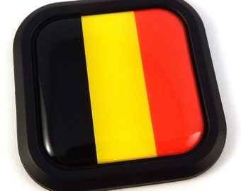 May include: A square emblem featuring the Belgian flag. The flag design includes vertical stripes of black, yellow, and red, encased in a black border. The emblem has a glossy finish.