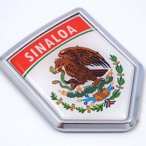 May include: A chrome-framed emblem featuring the Sinaloa state seal. The seal includes a brown eagle, a green snake, and a cactus, set against a white background. The word "SINALOA" is displayed in white letters on a red banner.