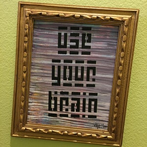 Puede incluir: Una obra de arte enmarcada con las palabras "USE YOUR BRAIN" en letras negras. El fondo tiene pinceladas horizontales coloridas. El marco es dorado y ornamentado, con una superficie texturizada.