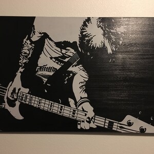 May include: Black and white painting of a musician playing a bass guitar. The artwork features a grayscale illustration of a person with long hair, wearing a jacket, and playing the instrument. The painting is signed in the bottom left corner.