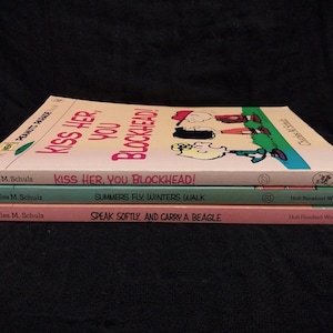 May include: Three vintage Peanuts books by Charles M. Schulz are stacked on a dark surface. The titles include "Kiss Her, You Blockhead!", "Summers Fly, Winters Walk", and "Speak Softly, and Carry a Beagle."