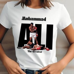 May include: White t-shirt with a graphic of Muhammad Ali in a boxing pose. The word "Muhammad" is above the word "ALI" in large black letters. The phrase "The Greatest" is below the image.