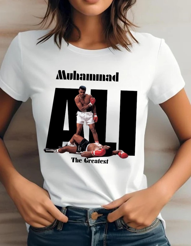 May include: White t-shirt with a graphic of Muhammad Ali in a boxing pose. The word "Muhammad" is above the word "ALI" in large black letters. The phrase "The Greatest" is below the image.