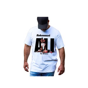May include: White t-shirt with a graphic of Muhammad Ali in a boxing match. The word "Muhammad" is above the word "ALI" in large black letters. The phrase "The Greatest" is below the image.