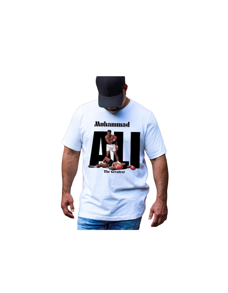 May include: White t-shirt with a graphic of Muhammad Ali in a boxing match. The word "Muhammad" is above the word "ALI" in large black letters. The phrase "The Greatest" is below the image.