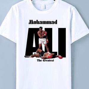 May include: White t-shirt with a graphic of Muhammad Ali standing over a defeated boxer. The text "Muhammad Ali" is above the image, with "The Greatest" below. The image shows Ali in boxing attire.