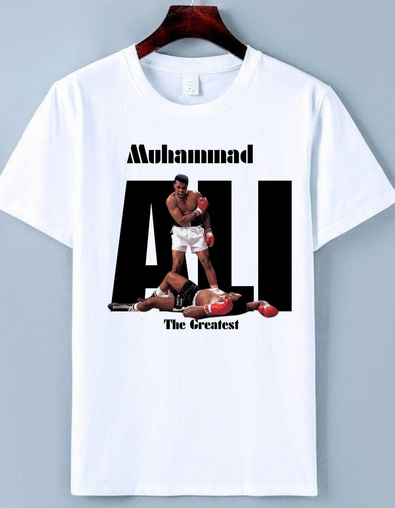 May include: White t-shirt with a graphic of Muhammad Ali standing over a defeated boxer. The text "Muhammad Ali" is above the image, with "The Greatest" below. The image shows Ali in boxing attire.