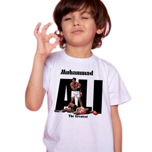 May include: White t-shirt with a graphic of Muhammad Ali in a boxing match. The text "Muhammad ALI The Greatest" is printed in black. The image is on a white t-shirt.
