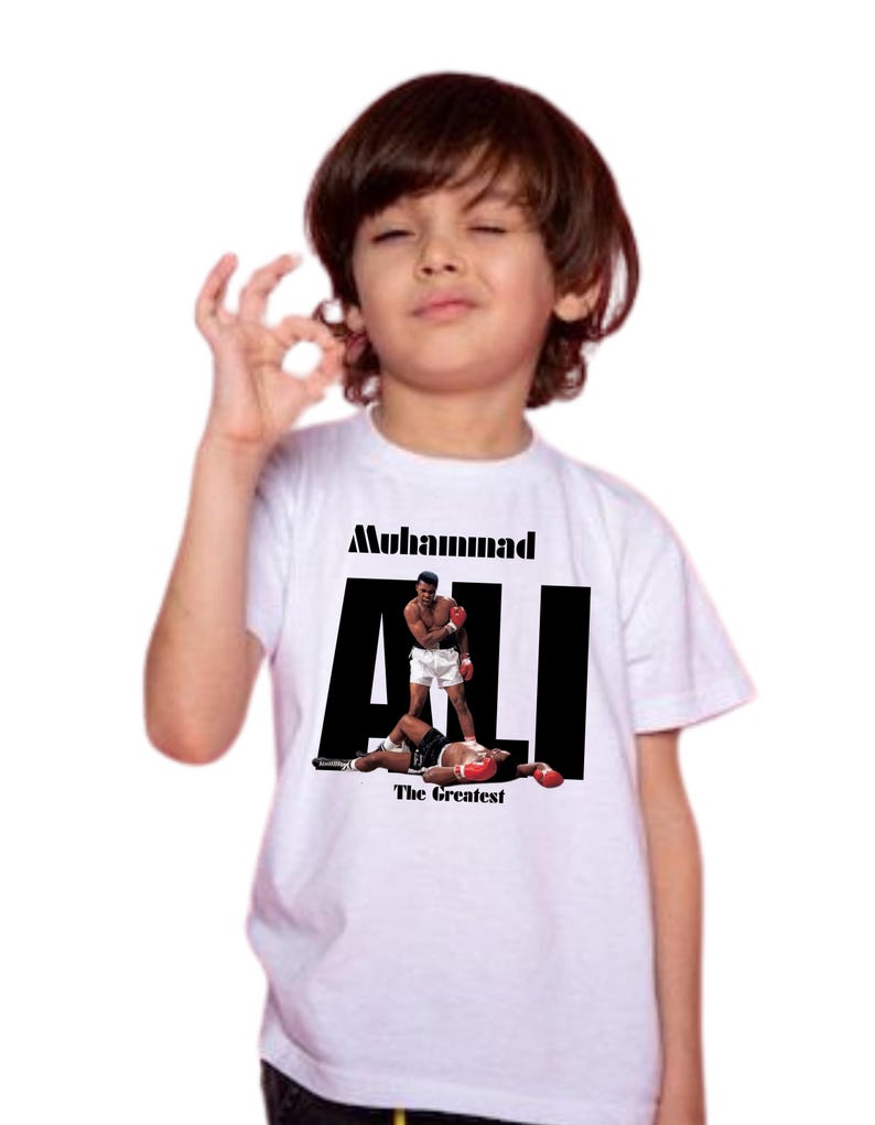 May include: White t-shirt with a graphic of Muhammad Ali in a boxing match. The text "Muhammad ALI The Greatest" is printed in black. The image is on a white t-shirt.
