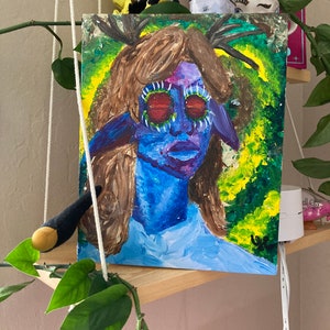 May include: A painting of a blue-skinned person with large, red-rimmed eyes and antlers. The person is wearing a light blue shirt and is surrounded by green and yellow foliage.