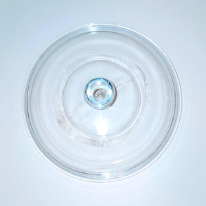 May include: A clear glass lid with a small, round knob in the center. The lid is circular with a slightly raised edge and a smooth, transparent surface. The knob has a blue-tinted appearance.