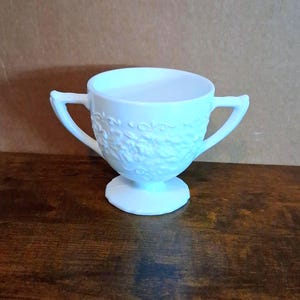 May include: A white, two-handled cup with a pedestal base. The cup features a floral relief pattern around the body and curved handles. The cup is made of a solid white material.