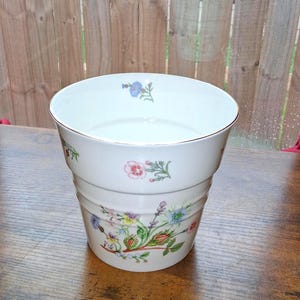May include: A white ceramic planter with a tapered shape and a gold-toned rim. The exterior features colorful botanical illustrations of various wildflowers and small blossoms scattered across the surface, with a larger floral arrangement at the base.