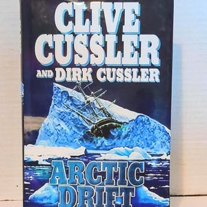 May include: A hardcover book titled "Arctic Drift" by Clive Cussler and Dirk Cussler. The cover features a ship caught in an iceberg, with the title in blue and white. The book is a Dirk Pitt novel.