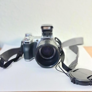 May include: A black and silver digital camera with a large lens, a pop-up flash unit, and a black neck strap. A circular black lens cap with the Sony brand name printed on it is resting on the surface next to the camera body and lens assembly.
