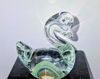 May include: A clear glass swan figurine with a curved neck and body. The sculpture has a green and white swirl design inside. The swan is displayed on a black surface.