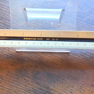 May include: A Staedtler Mars triangular scale ruler featuring a tan top edge with imperial markings up to 12 inches, a black center section with white text, and a light green bottom edge with metric markings up to 30 centimeters, made in Japan.