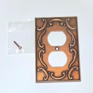 May include: A decorative copper-colored double outlet cover with an ornate scroll design. Includes a small screw and a clear plastic bag. The cover has two circular openings and a small central hole.