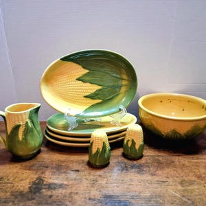 May include: A ceramic corn-themed dinnerware set. The set includes plates, a serving bowl, a pitcher, and salt and pepper shakers. The plates and serving bowl are yellow and green, with a corn design. The pitcher and shakers are also yellow and green.