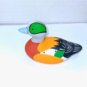 May include: A ceramic duck figurine with a white head, green eye patch, and blue beak. The body features orange, red, and black feathers with white accents. This decorative item is a colorful addition to any home decor.