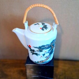 May include: White ceramic teapot with a woven handle and a lid. The teapot is decorated with a painted scene of a tree and birds in shades of blue, green, and red. The teapot is sitting on a black cube.