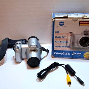 May include: A silver Konica Minolta DiMAGE Z10 digital camera with a black lens cap, strap, and remote. The camera is next to its original box, which features the camera's image and the text "32X" and "Rapid AF".