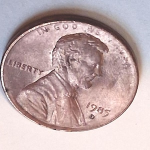 May include: A copper-colored United States one-cent coin featuring the profile of Abraham Lincoln facing right. The text LIBERTY is on the left, IN GOD WE TRUST is at the top, and the year 1985 with the mint mark D is on the right.