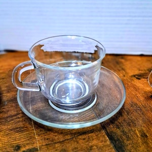 May include: A clear glass teacup and saucer set. The teacup has a rounded bowl and a curved handle. The saucer is a flat, circular plate. The set is made of transparent glass, allowing light to pass through.