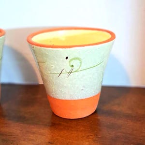 May include: A small, hand-painted ceramic pot with a light green exterior, orange base, and orange rim. The pot has a yellow interior and is decorated with a simple green swirl design and black lines. The pot is sitting on a wooden surface.