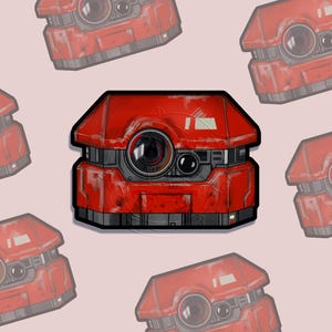 May include: A red, geometric robot sticker with a distressed, metallic finish. The robot has two large, circular lenses and a smaller lens. The sticker is surrounded by faded, repeating images of the same robot on a light pink background.