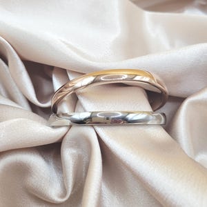 May include: Two smooth, polished metal bangles are stacked together. The top bangle features a warm rose gold tone, while the bottom bangle has a cool, reflective silver tone. Both bracelets have a rounded, solid profile with a high-shine finish.