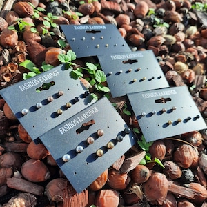 May include: Five black cards with the text "Fashion Earrings" on each card. Each card has a set of earrings with different colors and styles. The earrings are silver, gold, black, and rose gold.